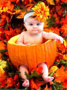 baby with a headband sits inside of a pumpkin with a fall scene behind her. 