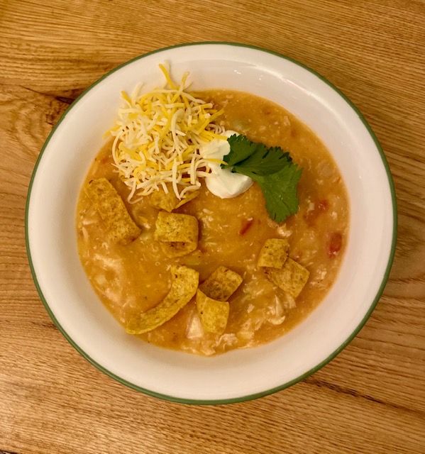 A white bowl sits on a wooden table.  The bowl is filled with a golden soup with red bits of tomato in it.  It is topped with a few sprigs of cilantro, a small amount of sour cream, shredded cheese and some Fritos are sprinkled on top as well. 