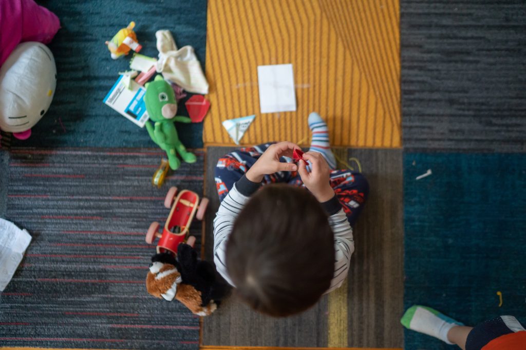 A young child sits on a carpeted floor playing with toys that are strewn around them. 