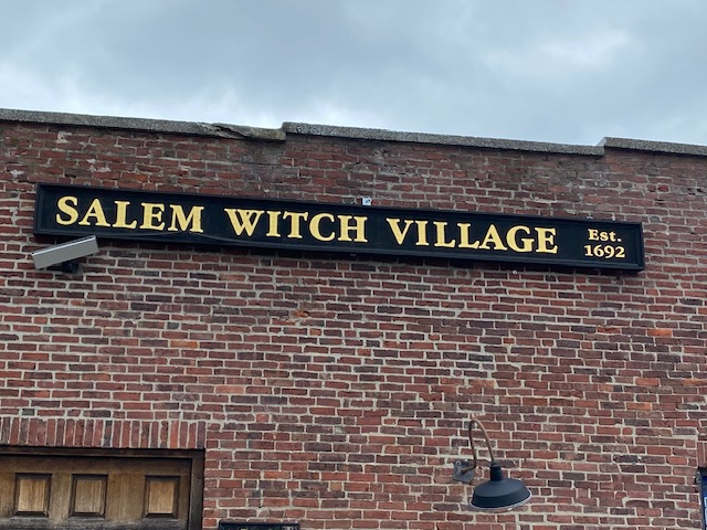 Old brick building with a large sign.  Sign is black with gold lettering that says Salem Witch Village.