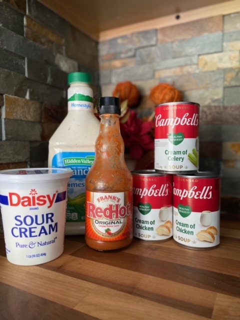 A photo from left to right of, Daisy sour cream, a large bottle of Hidden Valley Ranch dressing, a bottle of Frank's Red Hot sauce and stacked in a pyramid, 2 cans of Campbell's cream of chicken soup and a can of cream of celery.  All items are on a wooden counter top with grey/brown brick behind them as well as a small orange floral arrangement. 