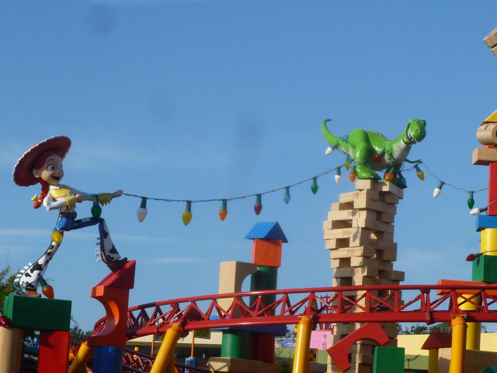 Photo of Toy Story Land in Hollywood Studios Disney World. A giant Jessie from the movie Toy Story, along with Rex the dinosaur string Christmas lights in "Andy's backyard" amongst giagantic toy blocks, car track (which is also a roller coaster track) and Jenga blocks.