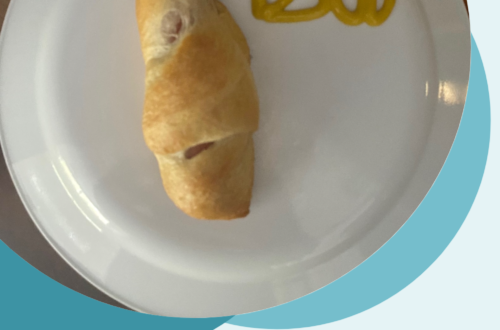 Picture of a hot dog mostly wrapped in a cooked crescent roll. The small bit of hot dog showing at the top has a mustard smiley face drawn on it. The word 'Boo' is also written to the right in mustard. This is all on top of a white plate, which is on top of a wooden counter top. It is surrounded with a light blue background. In the bottom right corner are the words, "Hot Dog Mummies!"