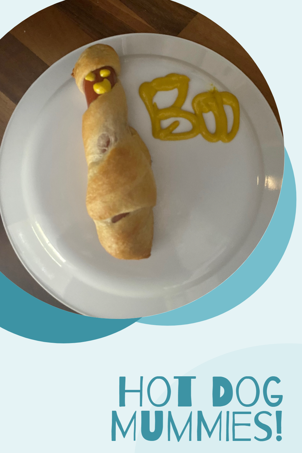 Picture of a hot dog mostly wrapped in a cooked crescent roll. The small bit of hot dog showing at the top has a mustard smiley face drawn on it. The word 'Boo' is also written to the right in mustard. This is all on top of a white plate, which is on top of a wooden counter top. It is surrounded with a light blue background. In the bottom right corner are the words, "Hot Dog Mummies!"