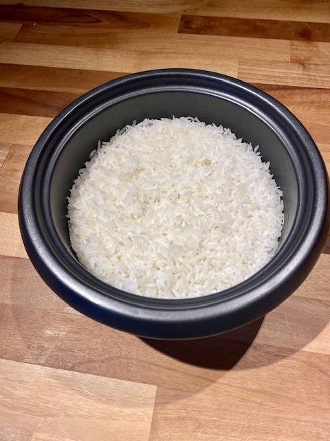 Cooked white rice inside of a black rice cooker. The cooker is placed on top of a wooden counter top.