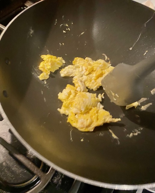 Scrambled eggs cook in a black wok. A grey rubber spatula is shown.