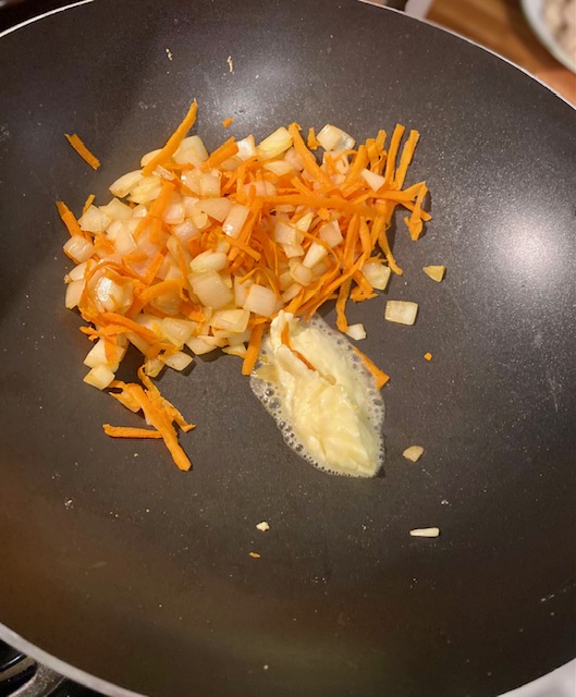 A mixture of chopped white onion and shredded carrot is to the upper left of a black wok. Garlic butter melts below the vegetables.