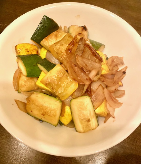 Browned pieces of yellow squash, zucchini and slices of onion in a white bowl.