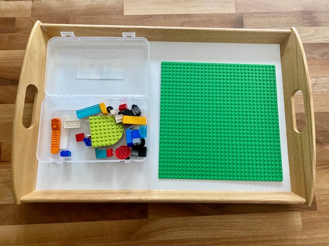 A wooden tray with a white bottom sits on a wooden counter top. Inside the tray on the left is a clear pencil box, opened with various Lego bricks inside.  To the right is a green Lego base plate.  