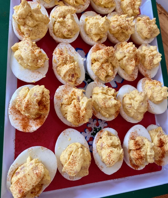 Over a dozen deviled eggs sit on a tray. The tray has a red bottom outlined with a white stripe. The rim of the tray is holiday green. Each deviled egg is filled with yellow filling and topped with red sprinkles of paprika.