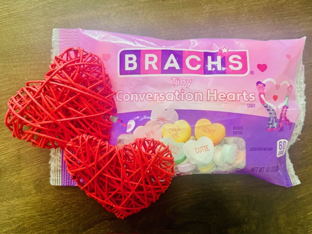a pink and purple bag of Valentine's Day Brach's Conversation Hearts sit on a brown table.  On top of the right corner of the bag are two red, wicker hearts about 3 inches tall each. 