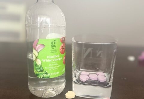 On a brown table, from left to right, sit a small clear bottle of white vinegar and short glass. The vinegar bottle has a bright green label. The glass is square and has five purple candy hearts at the bottom. In front of both items sits a small pile of multicolor candy hearts.