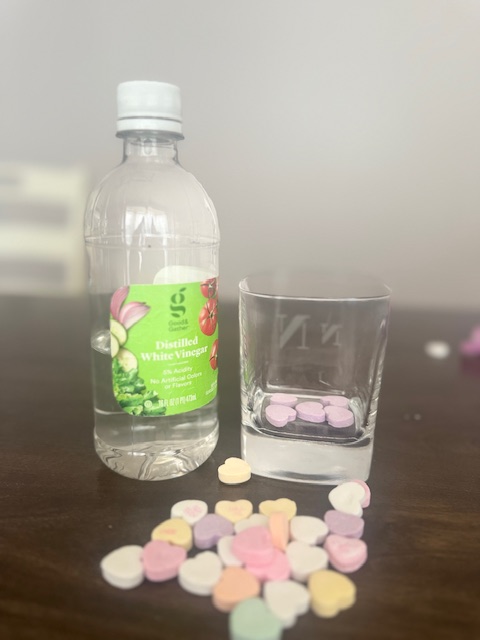 On a brown table, from left to right, sit a small clear bottle of white vinegar and short glass. The vinegar bottle has a bright green label. The glass is square and has five purple candy hearts at the bottom. In front of both items sits a small pile of multicolor candy hearts.
