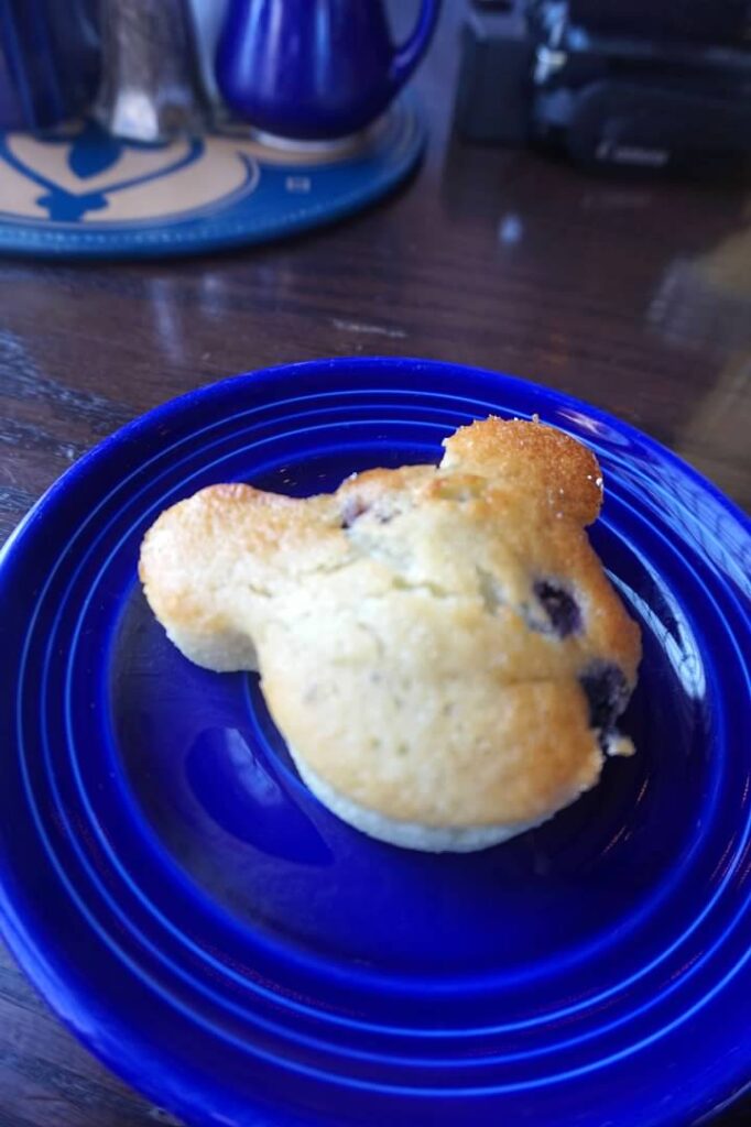 A blueberry muffin in the shape of Mickey Mouse. It is plated on a small royal blue plate which sits on a brown table. 