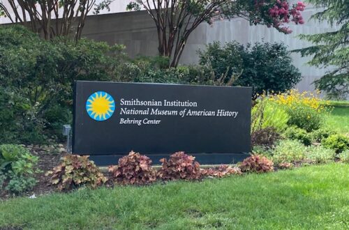 A large, rectangular, black sign for the Smithsonian museums is in a flower bed. The flowers are dark red. The sign is long and has the Smithsonian logo (a yellow sun with a bright blue circle around it) on the left side. In white lettering the sign reads, "Smithsonian Institution-National Museum of American History-Behring Center". The sign sits in front of various trees in a grassy area.