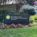 A large, rectangular, black sign for the Smithsonian museums is in a flower bed. The flowers are dark red. The sign is long and has the Smithsonian logo (a yellow sun with a bright blue circle around it) on the left side. In white lettering the sign reads, "Smithsonian Institution-National Museum of American History-Behring Center". The sign sits in front of various trees in a grassy area.