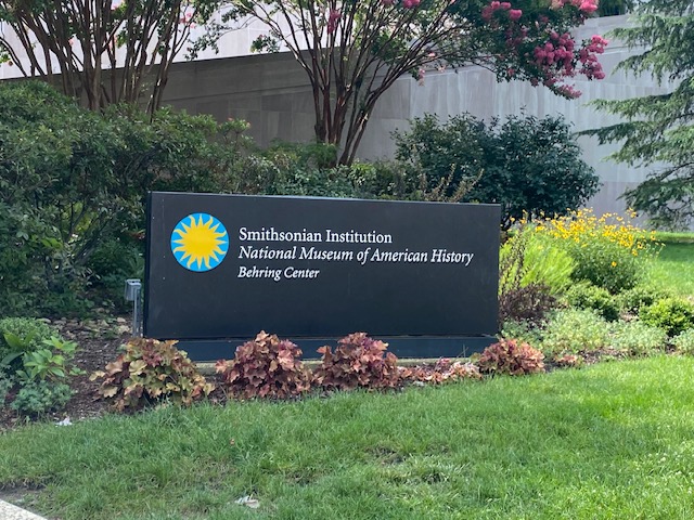 A large, rectangular, black sign for the Smithsonian museums is in a flower bed. The flowers are dark red. The sign is long and has the Smithsonian logo (a yellow sun with a bright blue circle around it) on the left side. In white lettering the sign reads, "Smithsonian Institution-National Museum of American History-Behring Center". The sign sits in front of various trees in a grassy area.