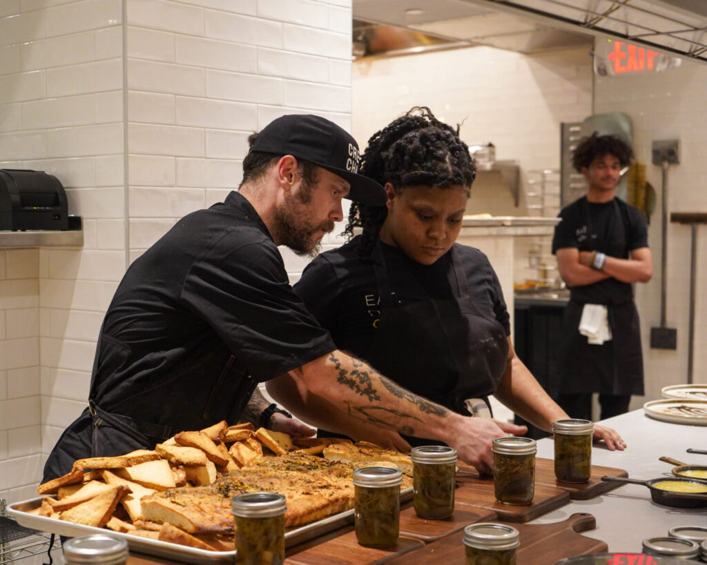 Two staff members are dressed in black chef coats.  The man on the left wears a hat, the woman on the right does not.  There are standing in a Cafe Momentum restaurant kitchen and prepping a large tray of food. It appears the food is slices of light brown bread, sliced in triangles.  There are mason jars of a dark substance lined up in front of the large silver baking sheet of bread.  A third staff member stands in the back right corner of the kitchen. 