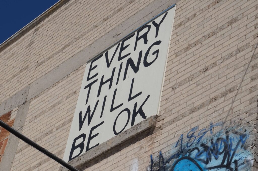 A large white rectangle is painted on the side of a tall brick building in Rapid City, South Dakota. The rectangle runs vertically.  In all capital, black letters read, "Every Thing Will Be Ok."  Some graffiti can be seen peeking up into the bottom righthand corner of the photo. 