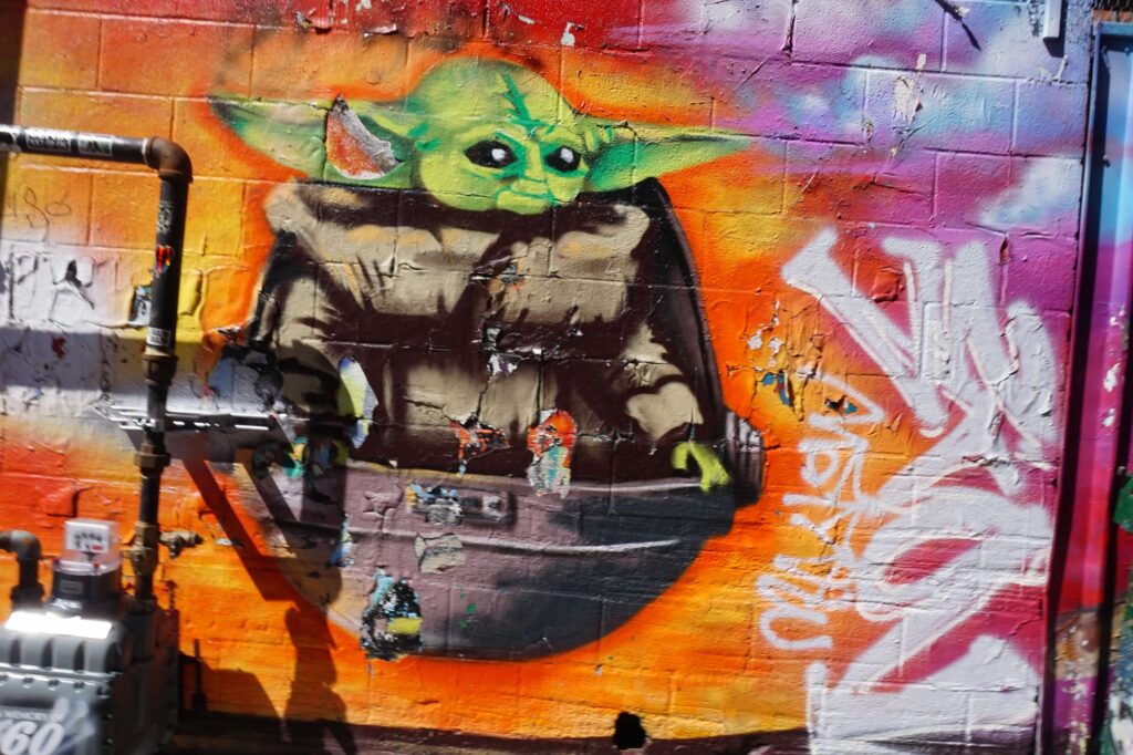 Art Alley in downtown Rapid City, South Dakota.  A graffiti style mural of Grogu (Baby Yoda from the Star Wars franchise) sitting in his "hovering pram". The background of the mural is bright orange, flowing into red towards the top.  Other white graffiti lettering can be seen on the right hand side of the photo. 