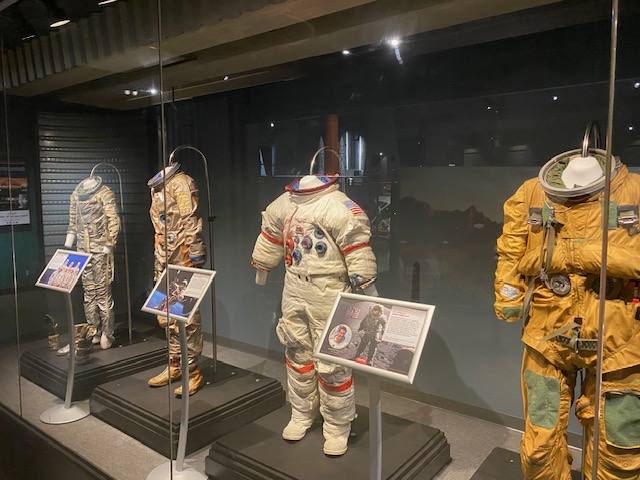 Behind glass at the U.S. Space and Rocket Center are pictured four space suits.  The suit on the right is yellow while the others are white.  There are museum display boards next to each suit.