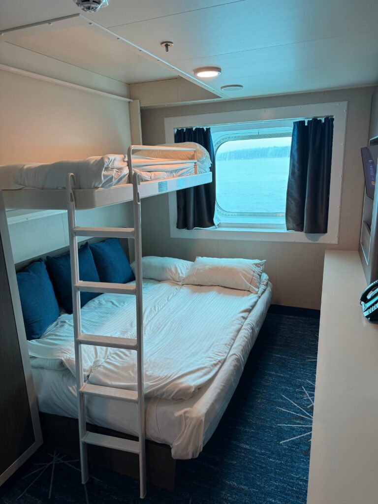 NCL Bliss Family Stateroom. Upper berth with a ladder down to the floor. Underneath the berth is a double bed.  A window is beyond both beds and a counter is to the left. 