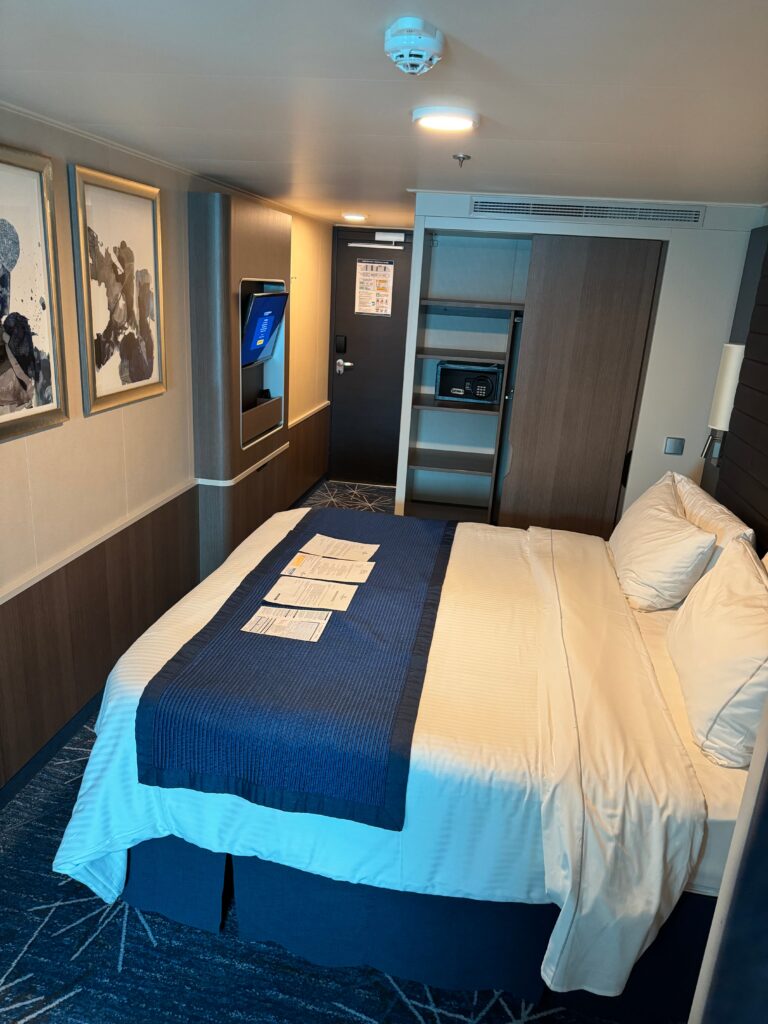 Family oceanview stateroom on the NCL Bliss  showing queen sized bed.  An open closet door shows a small safe.  Across from the bed is a tv and some artwork.  