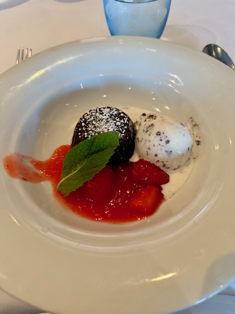 Dessert at Savor restaurant on the NCL Bliss- A round chocolate cake with powdered sugar sprinkled on top sits next to a scopp of chocolate chip ice cream. Below is a puddle of strawberries and sauce with a mint leaf on top.