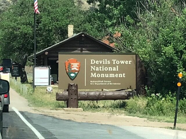 A brown sign reads: Devvils Tower National Monument in large cream colored letters. Beneath, in smaller letters reads: National Park Service/U.S. Department of the Interior
The wording is on the lefthand side of the sign with the orange and green National Parks logo on the right side.  The sign is the size of a small billboard.  Behind the sign is a small brown cabin, surrounded by a wooded area.  Part of the road is visibile on the front and left side of the photo. 