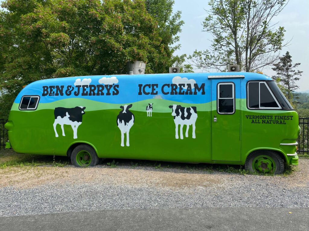 A large RV or bus is completely painted in a mural.  There is a blue sky and cows on grass.  On the sky reads, "Ben & Jerry's Ice Cream". In smaller letters underneath the front, side window reads, "Vermont's  Finest All Natural". 