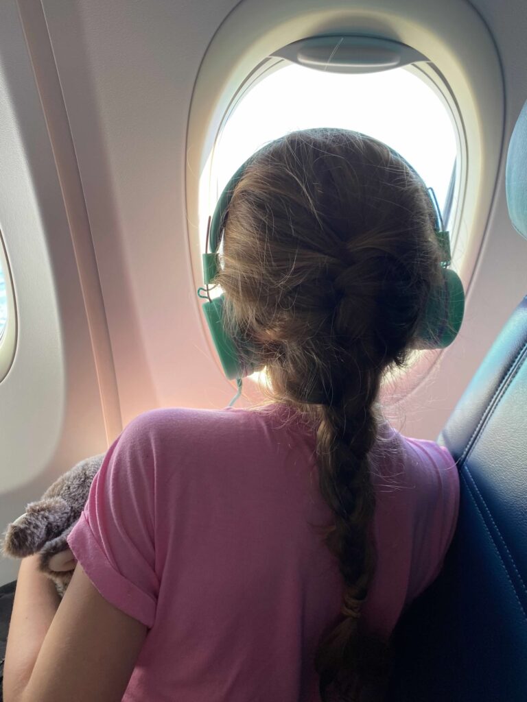 Travel via airplane. Pictured is the back of a young child. They have a long blonde braid and are wearing headphones. They are wearing a pink t-shirt. They look out an airplane window.