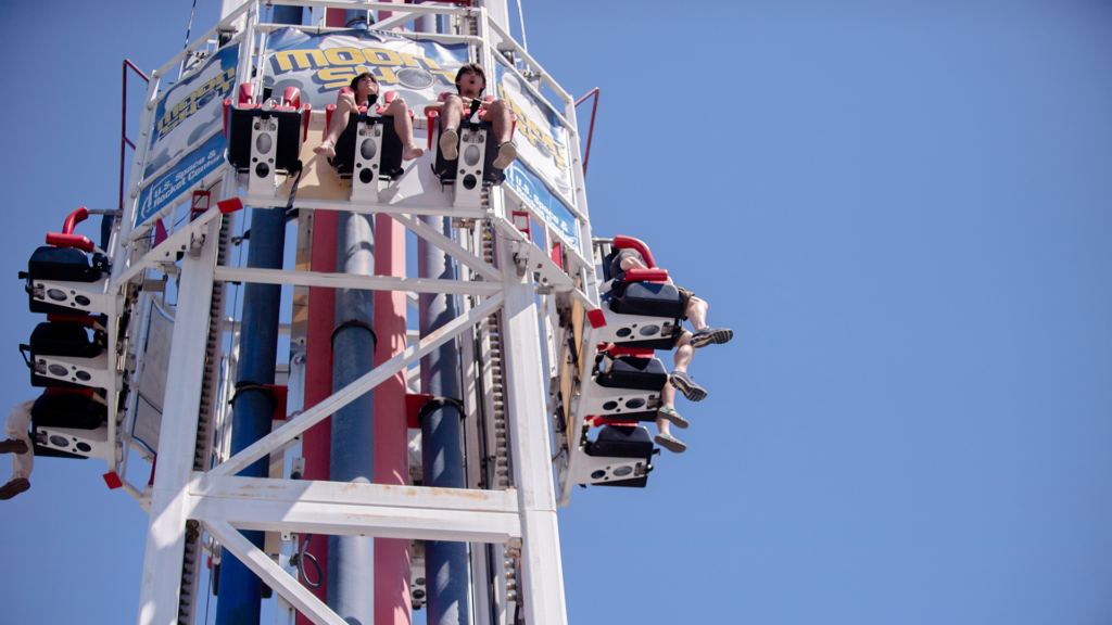 An amusement park type ride.  The ride is very tall and has a white structure.  Riders sit 3 across with black over the shoulder harnesses. The riders' legs dangle. The structure has four sides each featuring one row of riders.  Ride is outdoors on a sunny cloudless day at the U.S. Space and Rocket Center in Huntsville, Alabama.