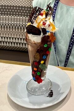 A chocolate milkshake has M&M candies stuck in chocolate on the side of the dish.  The shake has whipped cream, Oreo cookies, caramel and more M&M's on the top.  Available at Coco's, a specialty dining location on the NCL Bliss