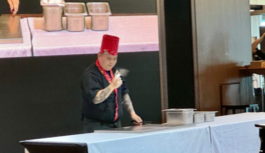 A Teppanyaki chef from the specialty dining restaurant on the NCL Bliss.  The chef wears a black chef coat with red trim and a red chef's hat. He is standing behind a long table and is tossing an egg so quickly that the egg is blurry. 