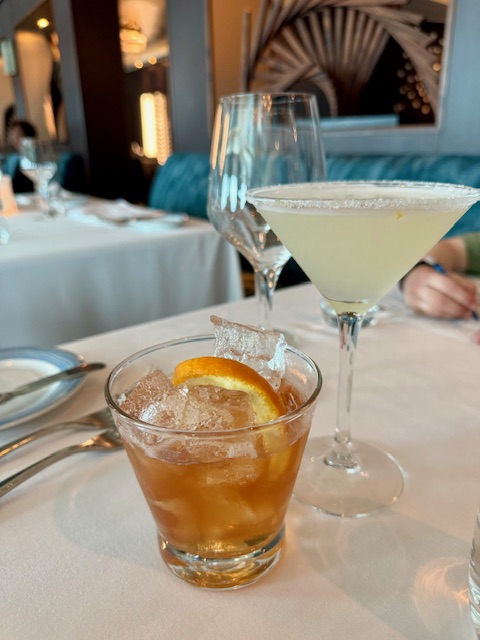 An Amber colored Old Fashioned with ice and an orange rind sits next to a martini glass with a pale yellow drink and a sugared rim.  The glasses are on a dining table with a white table cloth. 