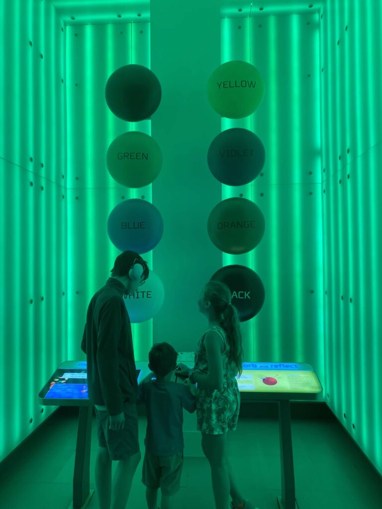 A tall room is cast in a green/turquoise light.  Circles in different colors and labelled with those colors are in two columns along the back wall.  Three children stand at a low podium with information about the exhibit at the Griffin Museum of Science and Industry in Chicago. 