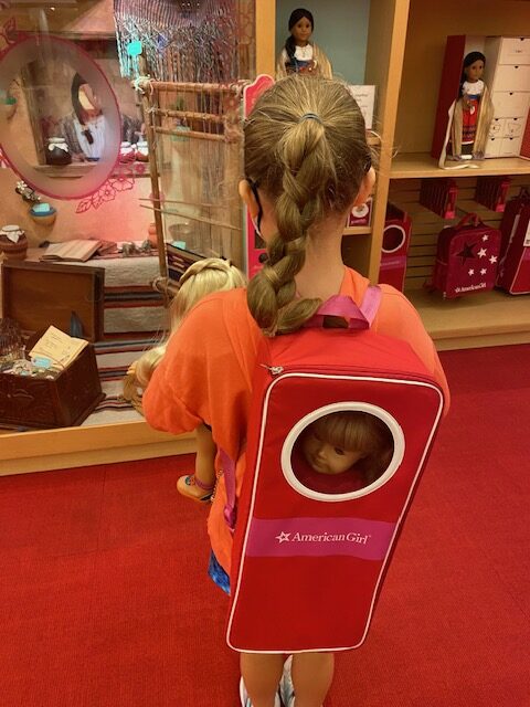 A girl shops in the American Girl store.  She wears a backpack that hold a doll and carries another doll in her arms. 