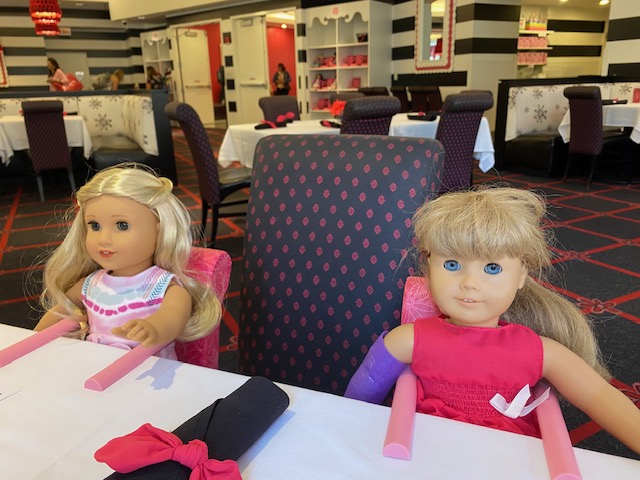 Two American Girl dolls sit in doll highchairs at a table in a restaurant.  There is a white tablecloth and black cloth napkins.