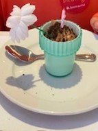 American Girl Cafe Chocolate Mousse served in a small blue pot. The pot looks like a flower pot and the mousse is covered in chocolate cookie crumbs to look like dirt.  There is a silk flower sticking out of it, as well as a birthday candle.  