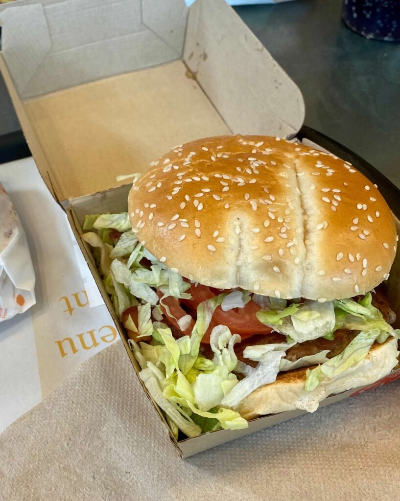 A photo of the 1955 Burger from the Chicago McDonalds location next to Hamburger University.  The burger is in a fast food burger box.  Theres is a sesame seed bun and a lot of shredded lettuce.  A tomato slice peeks out of the lettuce.  