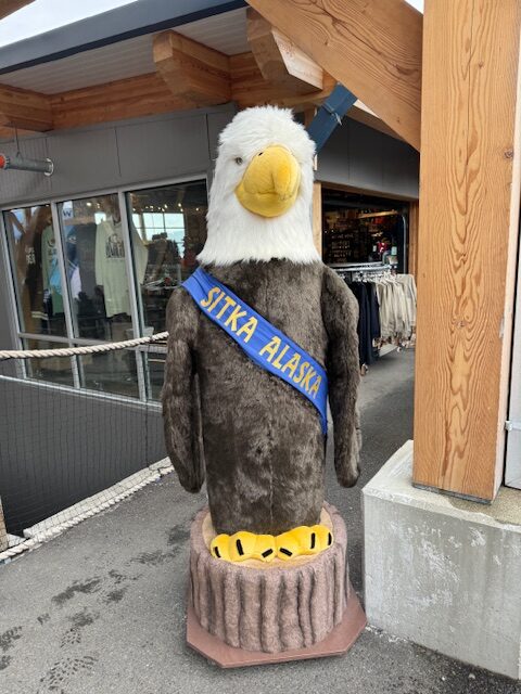 An approximately 6 foot tall stuffed eagle is outside of a building. It wears a blue sash that reads, "Sitka, Alaska".