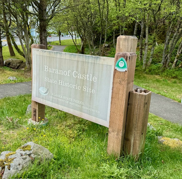 A large sign, weathered with streaks of rain, reads, "Baranoff Castle State Historic Site-Alaska State Parks Department of Natural Resources" in Sitka, Alaska