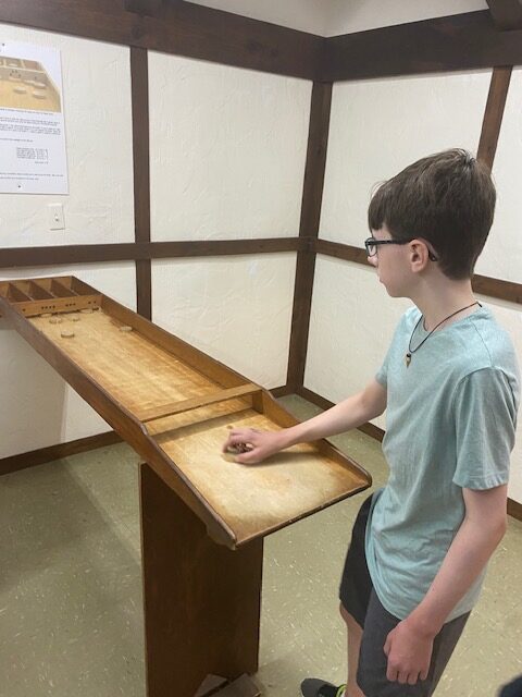 A child stands at a long wooden table. He holds a small wooden disc and attempts to get the disc in one of the slots at the end of the table.