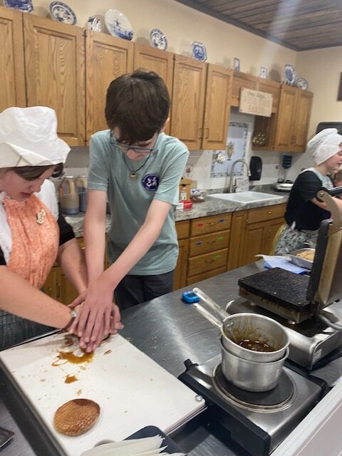 A child presses his hands on top of a cook's hands. The cook is dressed in traditional Dutch clothing as part of the Nelis' Dutch Village uniform. On the counter in front of them is a sauce pan and a waffle press. Their hands press down on a brown substance on top of a white cutting board. Behind them are oak kitchen cabinets.