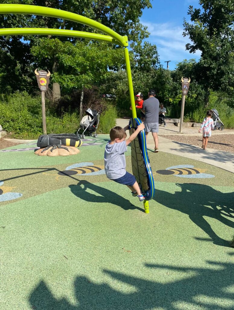 A child swings on a standing swing at Bison's Bluff playground in Schaumburg, IL. There is green rubber underneath and shadows are cast along the ground. The rubber flooring has bees printed on it and there is a large bumblebee for children to climb on. Other people and walking paths can be seen in the background.