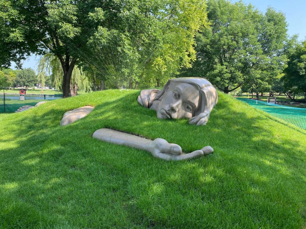 A large sculpture of a woman's head and hand come out of a green hill at the Chicago Atheneum Sculpture Park.