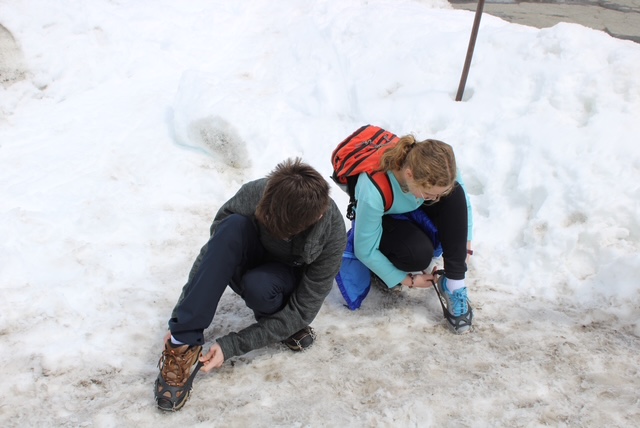 Two kids pull crampons onto their hiking shoes.  There is snow all around them. 