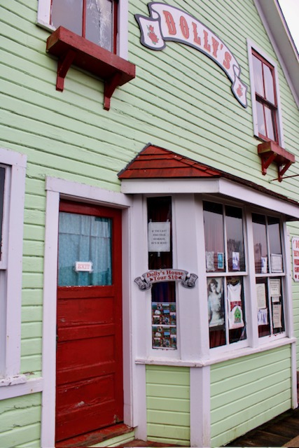 The side of a light green house.  The house has a red door and a sign that reads, "Dolly's" in red lettering on the second story.  The bay window on the first floor is filled with notices and signs from Ketchikan. 