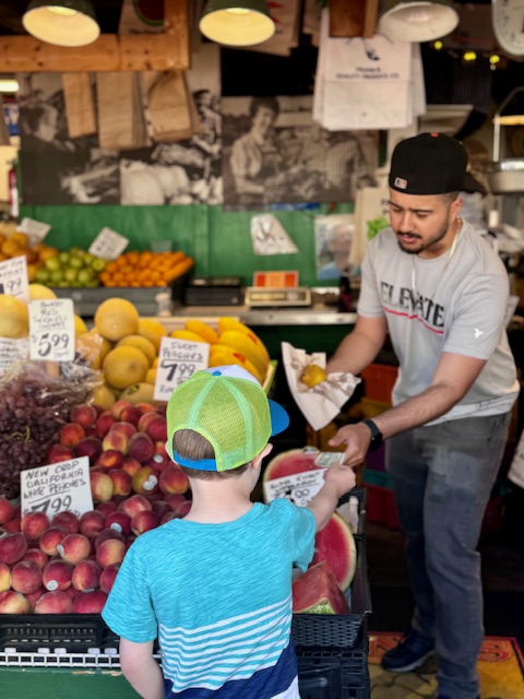 A child hands money to a stall owner at a fruit stand at Pike Place Market Seattle.  