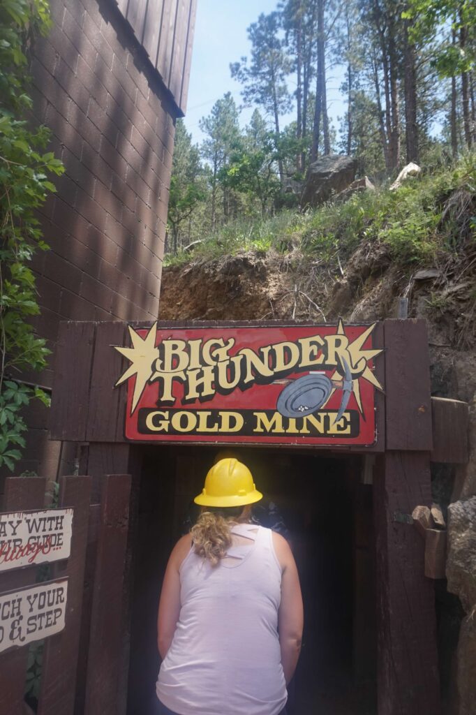 A person with a yellow hardhat bends down slightly to enter a tunnel to a gold mine.  A red Big Thunder Gold Mine sign hangs above the entrance. 